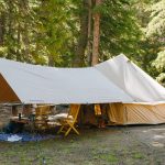 A large canvas bell tent with a sunshade tarp setup in a forest campsite, featuring chairs and camping gear under the canopy.