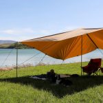 A large sunshade tarp set up by a lakeside, providing shade for two camping chairs and a person relaxing on the grass with a dog under it.