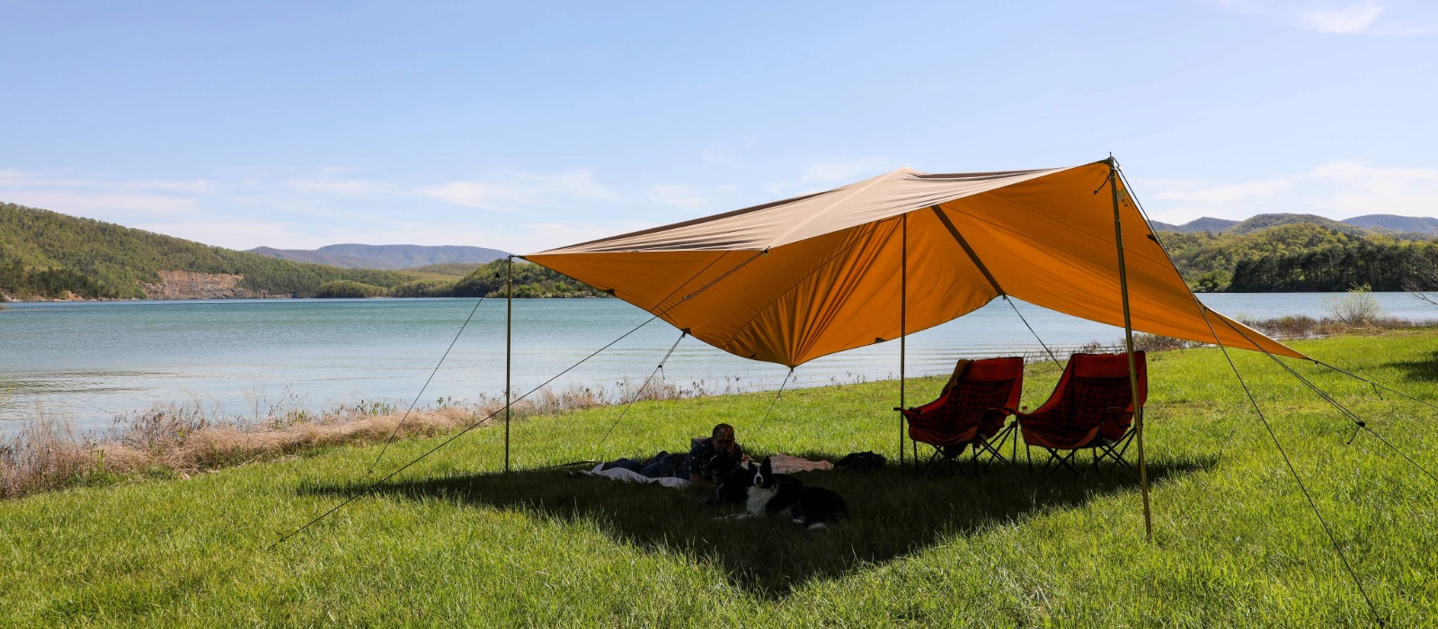 A large sunshade tarp set up by a lakeside, providing shade for two camping chairs and a person relaxing on the grass with a dog under it.