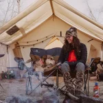 Woman sitting with her dog on chairs outside a wall tent porch, enjoying a cozy camping moment. Image credit: White Duck Outdoors.