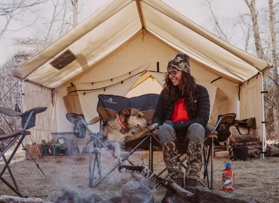 Woman sitting with her dog on chairs outside a wall tent porch, enjoying a cozy camping moment. Image credit: White Duck Outdoors.