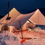 A warm canvas tent with a wood stove glowing at night, surrounded by snow and mountain views.
