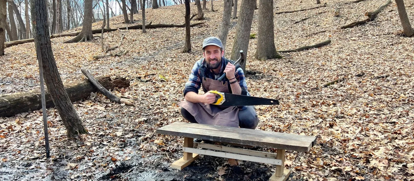 Person building a DIY wooden bench in a forest using hand tools and wearing a canvas apron.