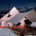 Insulated yurt tent with a stove and tarp in a snowy mountain landscape for cozy winter camping.