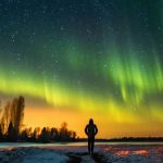 A person standing by a snowy riverside watching the northern lights glow across the night sky
