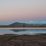 A calm lake reflecting distant mountains and a soft pastel sunset sky in a quiet open landscape.