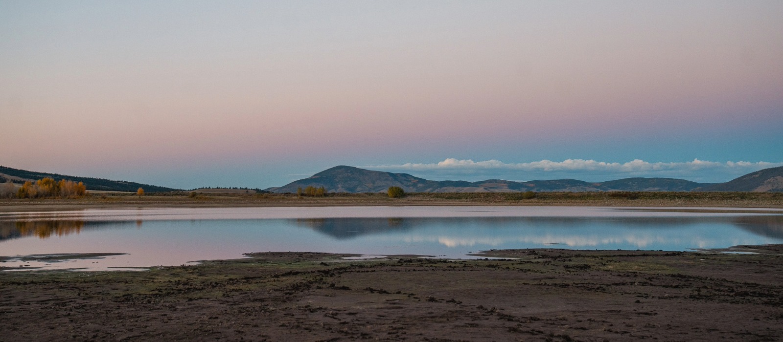 A calm lake reflecting distant mountains and a soft pastel sunset sky in a quiet open landscape.