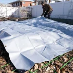 A person laying a poly tarp over a backyard garden bed to protect the soil during winter.