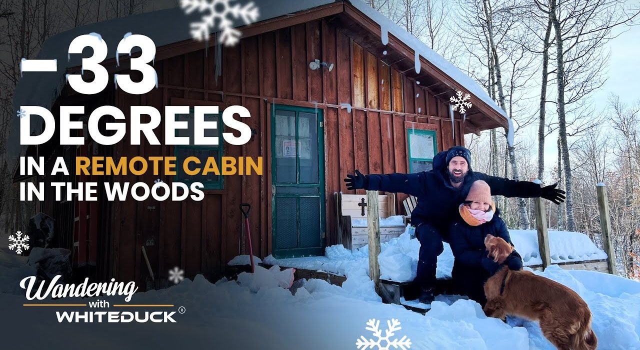 Couple sitting with their dog in deep snow outside a remote wooden cabin in the woods during extreme winter temperatures of minus 33 degrees.