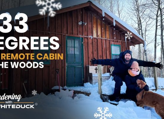 Couple sitting with their dog in deep snow outside a remote wooden cabin in the woods during extreme winter temperatures of minus 33 degrees.