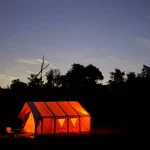 A glowing orange tent set up in a dark outdoor camp at dusk with a clear sky full of stars, perfect for a stargazing night.