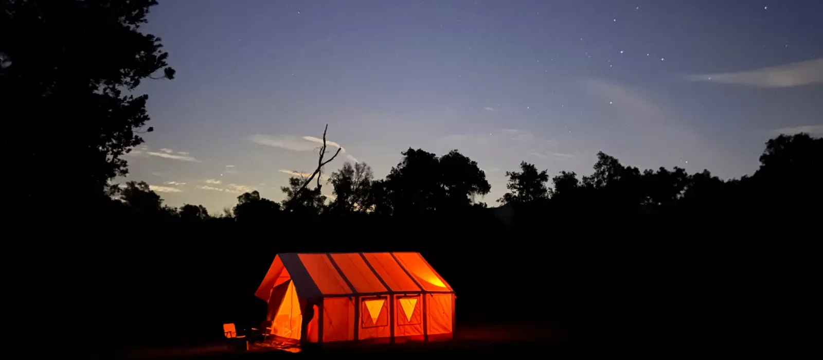 A glowing orange tent set up in a dark outdoor camp at dusk with a clear sky full of stars, perfect for a stargazing night.