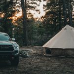 Toyota pickup truck parked beside a canvas bell tent in a forest campsite at sunset.