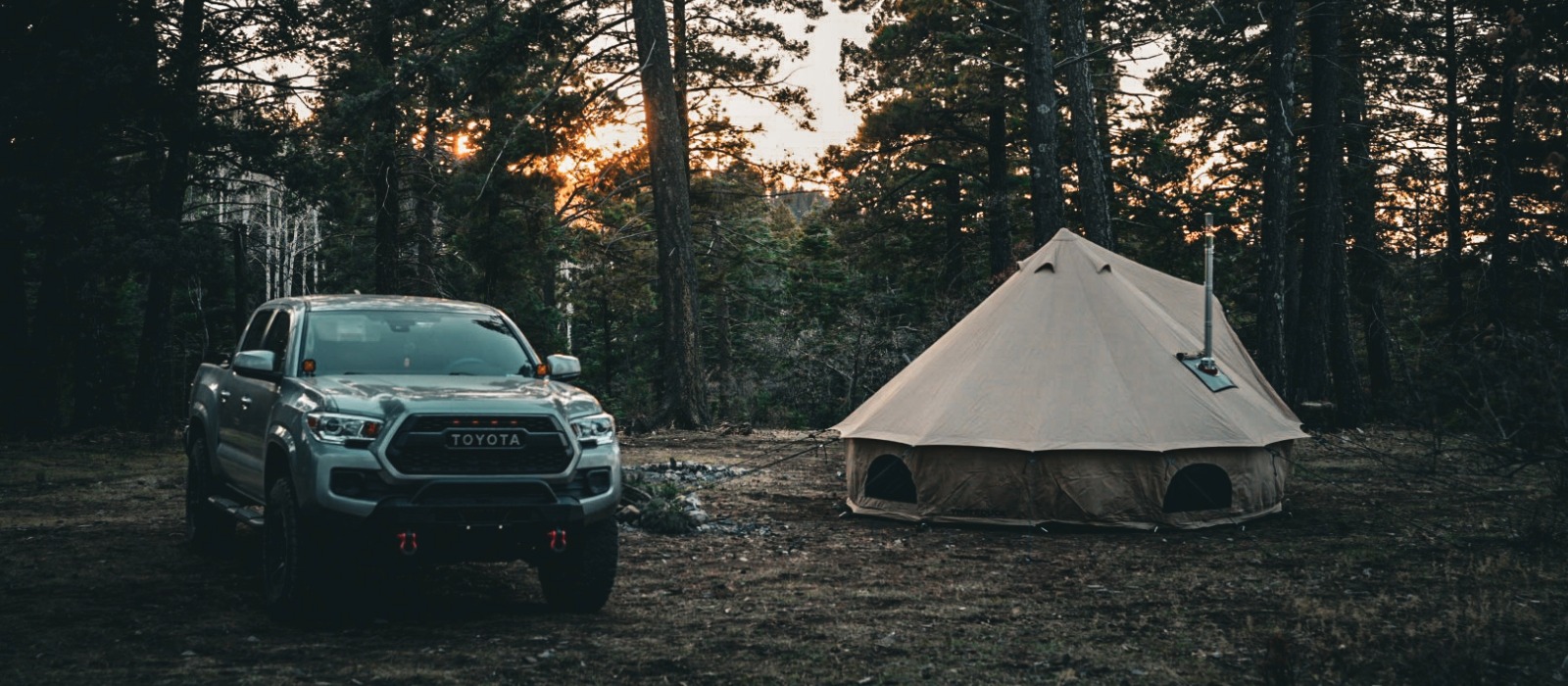 Toyota pickup truck parked beside a canvas bell tent in a forest campsite at sunset.