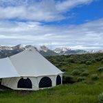 A large yurt tent set up in a green field with a mountain range in the background under a partly cloudy sky.