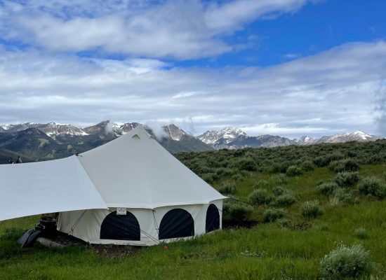 A large yurt tent set up in a green field with a mountain range in the background under a partly cloudy sky.