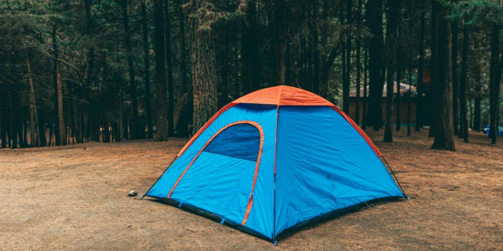 A blue pop-up tent with an orange rainfly set up in a pine forest clearing.