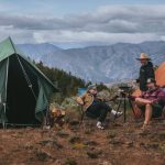 Friends enjoying camp cooking at an outdoor campsite, relaxing between canvas tents with mountains in the background