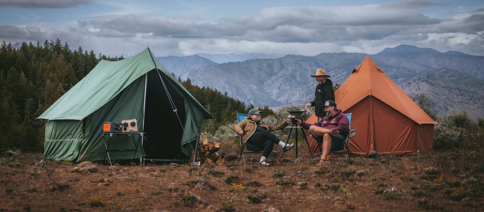 Friends enjoying camp cooking at an outdoor campsite, relaxing between canvas tents with mountains in the background
