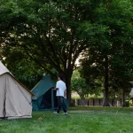 A peaceful backyard camping scene with multiple canvas tents set up under large green trees, featuring a person walking near the tents during daylight.