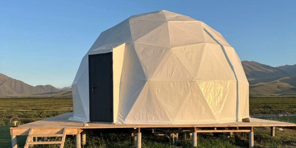 A white dome tent set up on a deck in a vast green field under a clear blue sky.