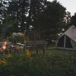 A canvas bell tent set up at a solo camping basecamp with a small campfire burning near outdoor seating surrounded by trees and greenery.