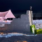 Winter camping scene featuring a canvas tent illuminated at night, surrounded by snow walls and blocks with a shovel placed nearby, showcasing a cold weather camping setup.