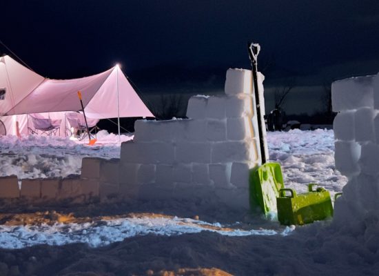 Winter camping scene featuring a canvas tent illuminated at night, surrounded by snow walls and blocks with a shovel placed nearby, showcasing a cold weather camping setup.