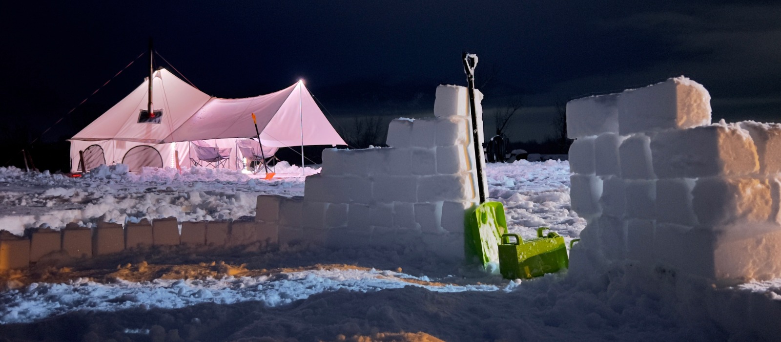 Winter camping scene featuring a canvas tent illuminated at night, surrounded by snow walls and blocks with a shovel placed nearby, showcasing a cold weather camping setup.