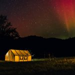 Hot tent camping at night with a glowing canvas tent under colorful northern lights in a forest clearing