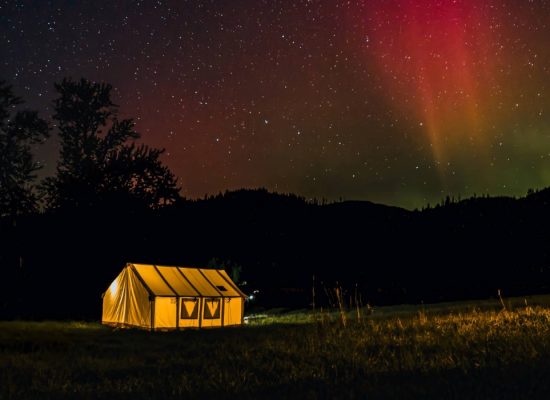 Hot tent camping at night with a glowing canvas tent under colorful northern lights in a forest clearing