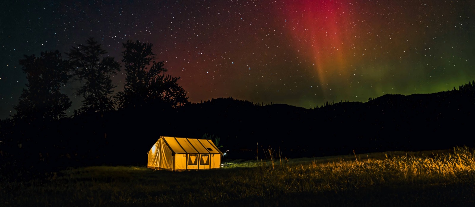 Hot tent camping at night with a glowing canvas tent under colorful northern lights in a forest clearing