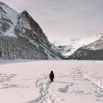A person walking across a frozen lake surrounded by snow-covered mountains and pine forests in a national park during winter.