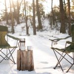 Two green camping chairs set in a snowy forest with a thermos and mugs on a tree stump, capturing a peaceful winter camping scene.