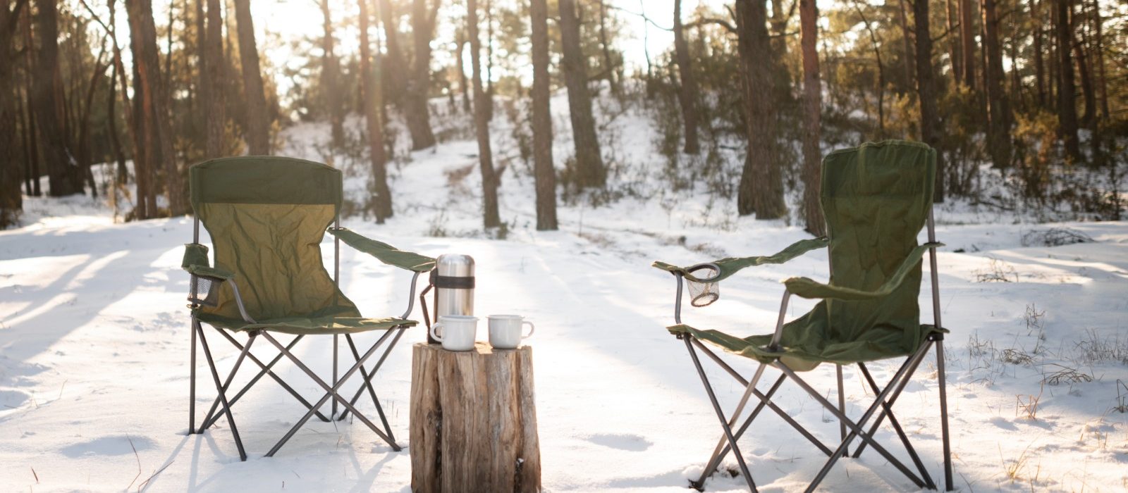 Two green camping chairs set in a snowy forest with a thermos and mugs on a tree stump, capturing a peaceful winter camping scene.
