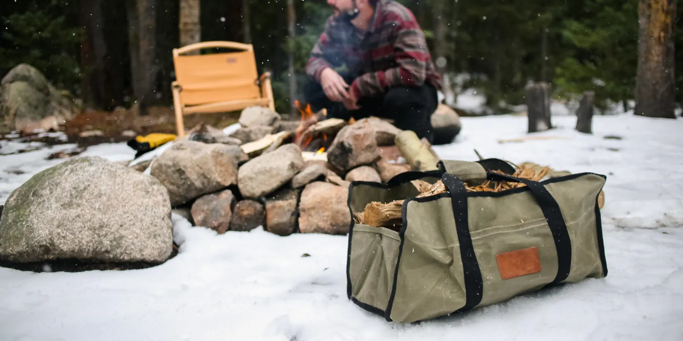 Canvas firewood carrier filled with logs at a snowy campsite.