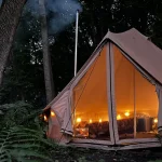A large canvas bell tent with a warm interior glow from string lights and a chimney pipe, pitched between mature trees in a forest during twilight