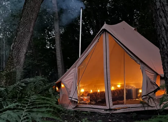 A large canvas bell tent with a warm interior glow from string lights and a chimney pipe, pitched between mature trees in a forest during twilight