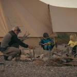 Man adding firewood to a stone fire pit while two young children sit in camping chairs nearby, in front of a large beige canvas bell tent with an awning at dusk.