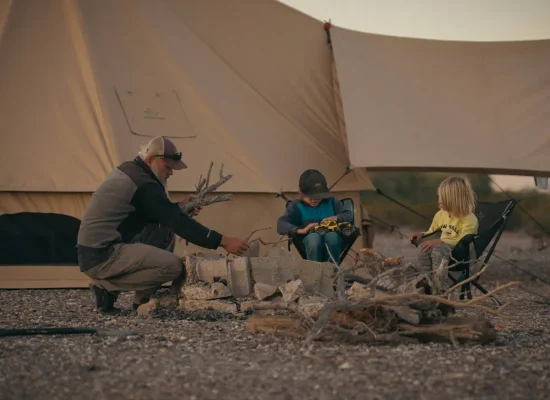 Man adding firewood to a stone fire pit while two young children sit in camping chairs nearby, in front of a large beige canvas bell tent with an awning at dusk.