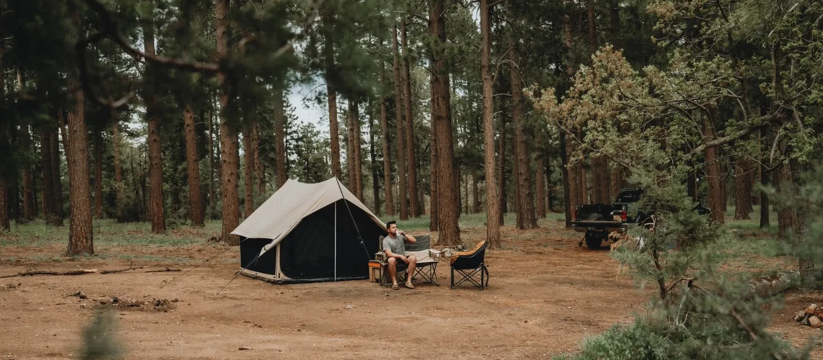 Man sitting in a camping chair, drinking a beverage outside a canvas Scout tent in a pine forest clearing, with a black pickup truck parked in the background.