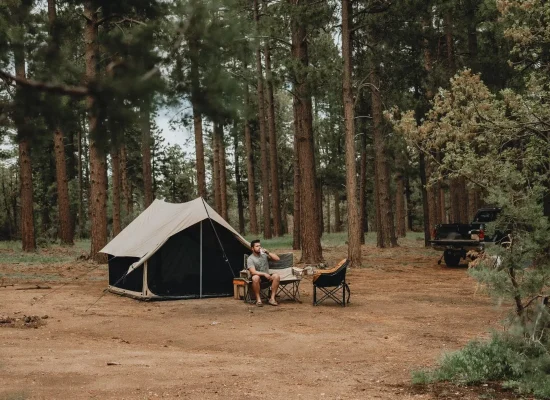 Man sitting in a camping chair, drinking a beverage outside a canvas Scout tent in a pine forest clearing, with a black pickup truck parked in the background.