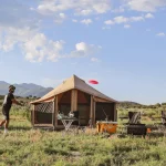 An outdoor campsite with canvas bell tents, a portable grill, and chairs, where two campers are enjoying a game of frisbee in a field with distant mountains.