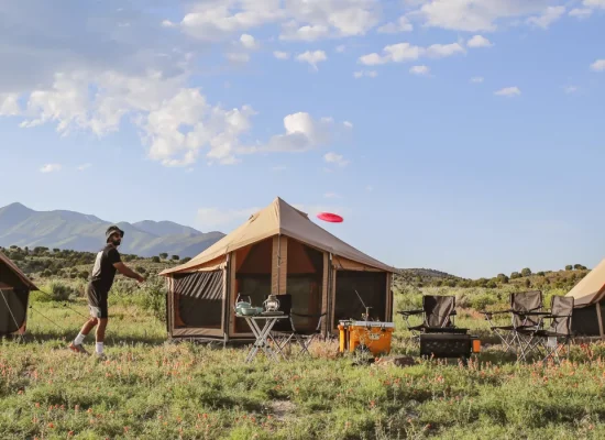 An outdoor campsite with canvas bell tents, a portable grill, and chairs, where two campers are enjoying a game of frisbee in a field with distant mountains.