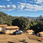 Canvas tents at a scenic hillside campsite with mountain views, trees, and parked vehicles under a blue sky.