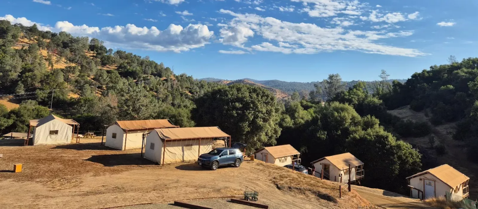Canvas tents at a scenic hillside campsite with mountain views, trees, and parked vehicles under a blue sky.