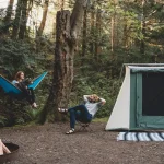 A couple relaxing at a forest campsite with their dog, featuring a blue hammock, a canvas tent, and a campfire setup.