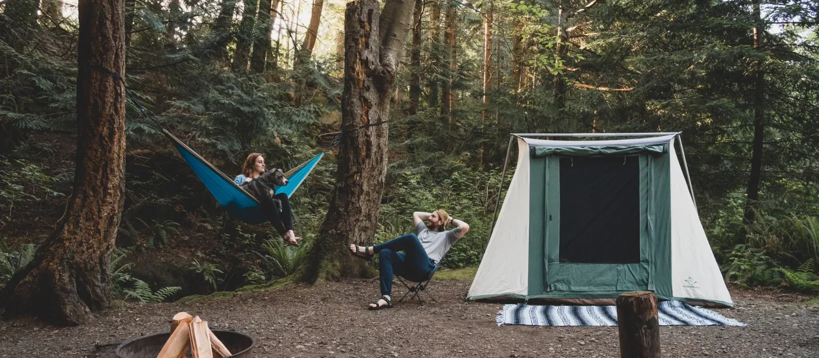 A couple relaxing at a forest campsite with their dog, featuring a blue hammock, a canvas tent, and a campfire setup.
