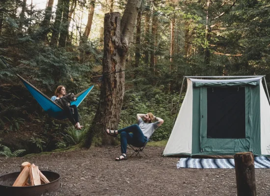 A couple relaxing at a forest campsite with their dog, featuring a blue hammock, a canvas tent, and a campfire setup.