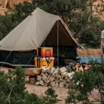 A man standing at a campsite nestled against rocky canyon walls, featuring a large tent, a neat pile of firewood, and a cooking station.